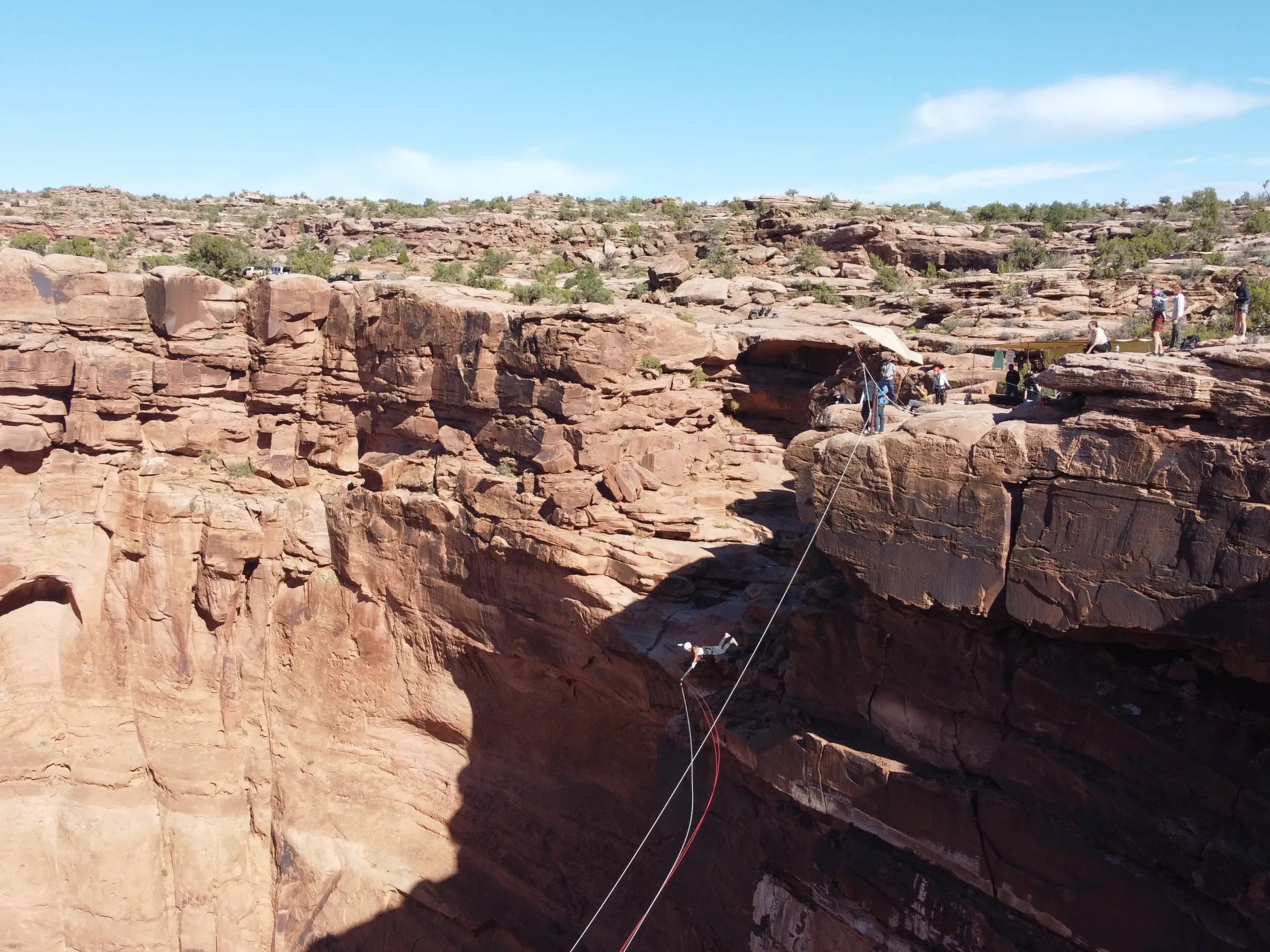 Person swinging on a rope between cliffs in a desert canyon with onlookers nearby.