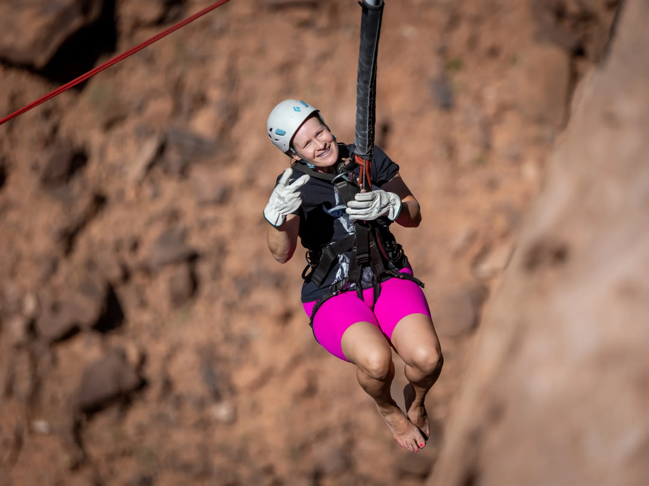 Person in pink shorts ziplining with a helmet and gloves against a rocky background.
