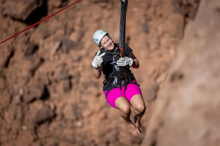 Person in pink shorts ziplining with a helmet and gloves against a rocky background.