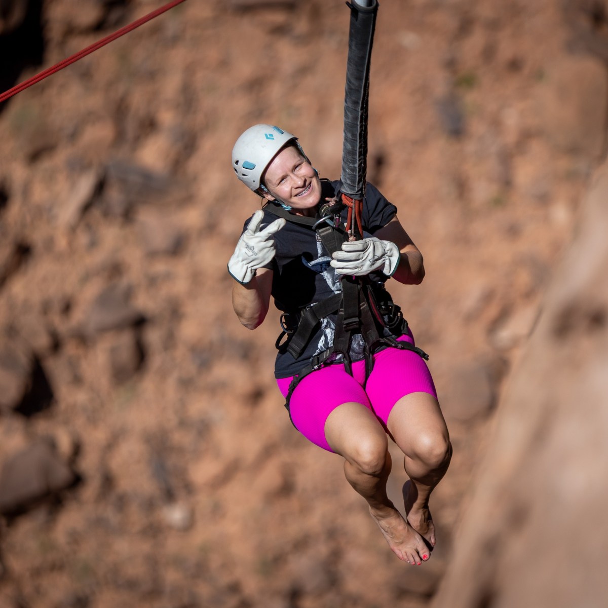 Person in pink shorts ziplining with a helmet and gloves against a rocky background.
