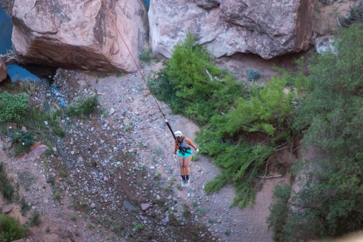Person in helmet swinging on rope over a rocky canyon with trees.