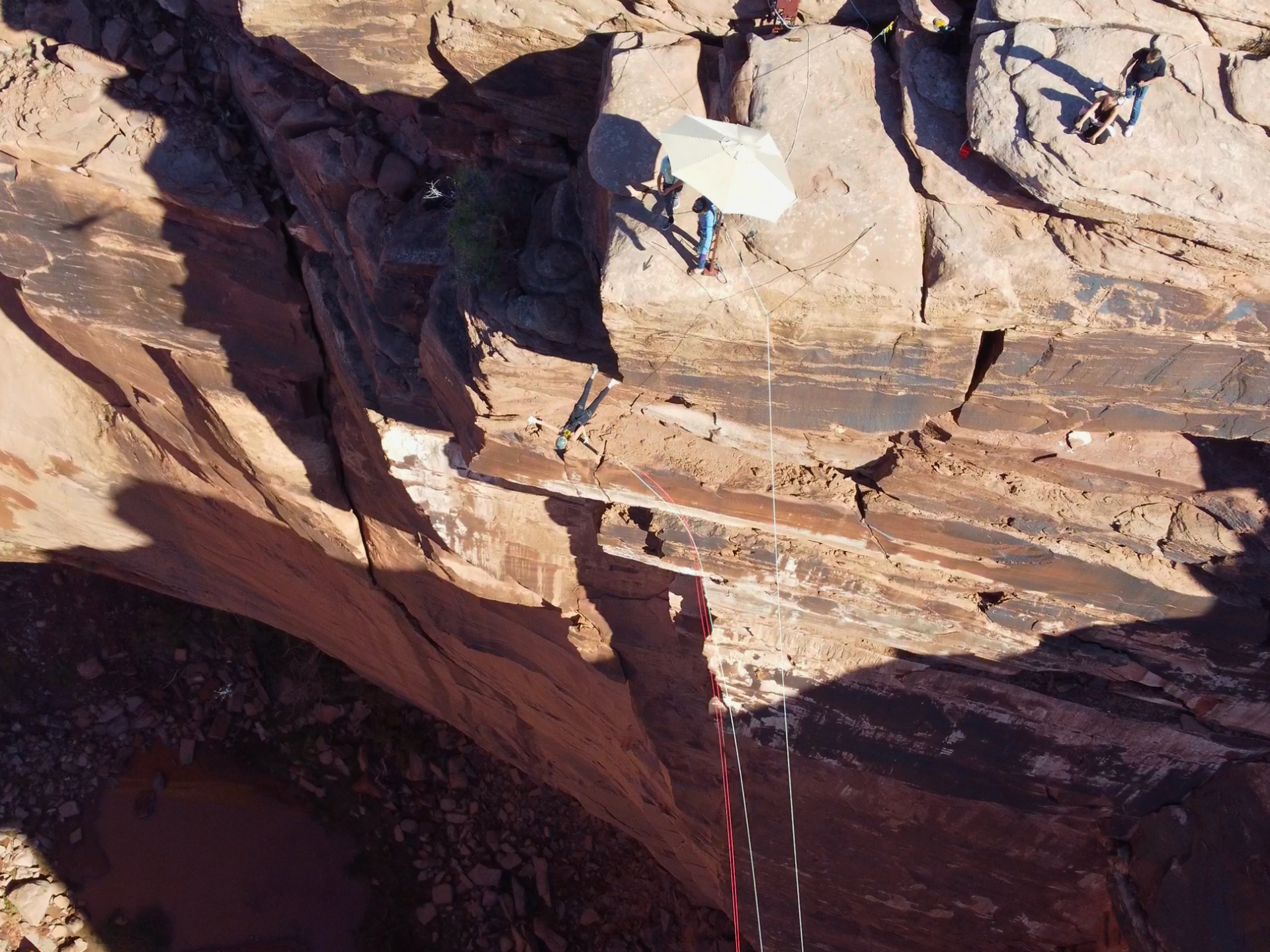 Two people on a rocky cliff with ropes and an umbrella, overlooking a canyon.