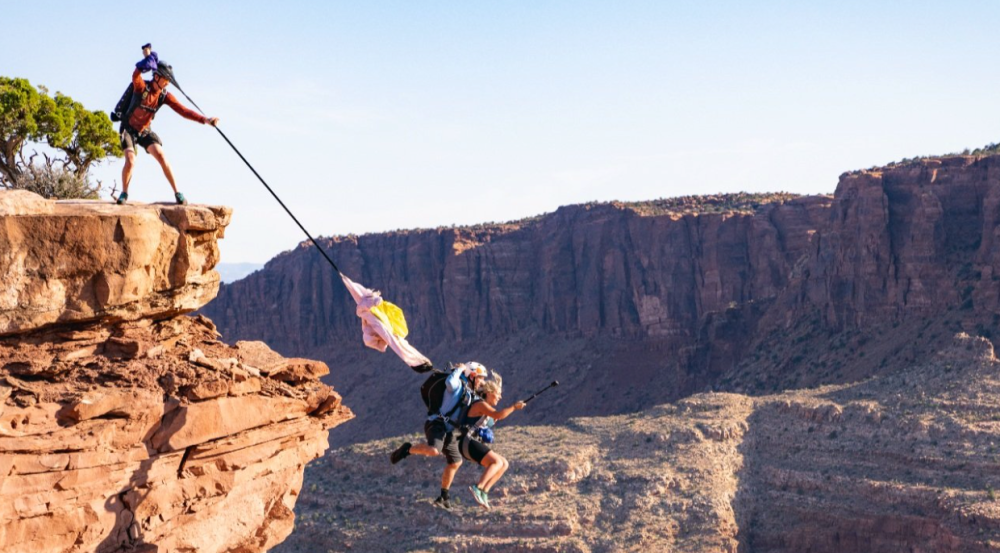Person jumps off cliff holding rope over canyon with another person standing at edge.