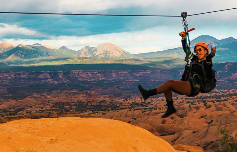 Person zip-lining over a canyon with mountains in the background, wearing a helmet and harness.