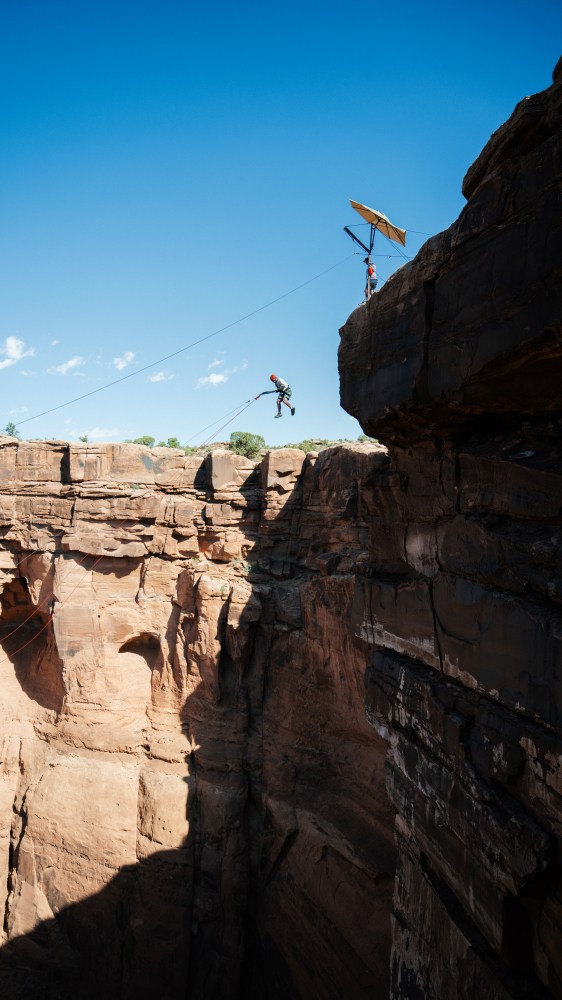 Person mid-jump across a canyon with safety harness attached to a line above.